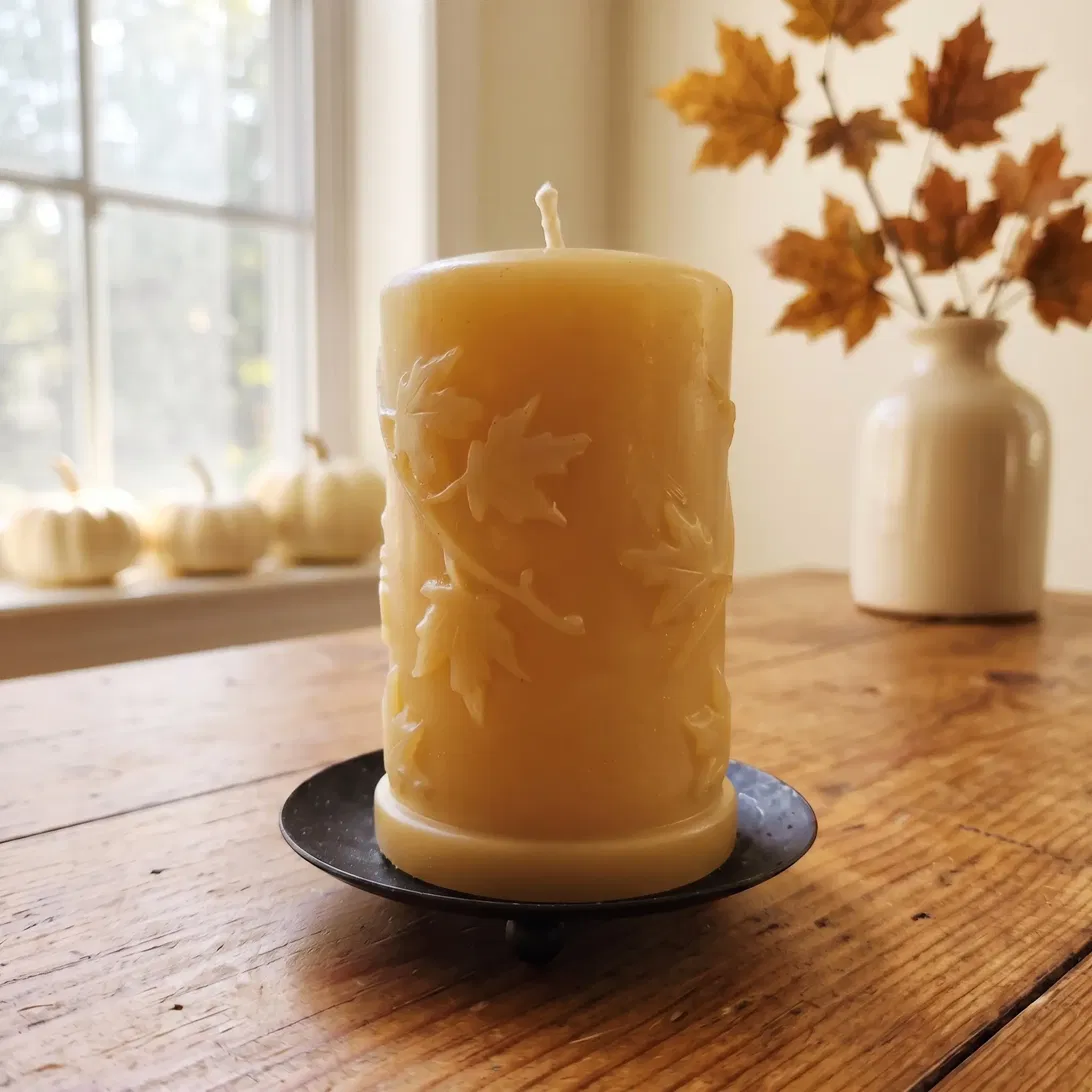 Autumn Leaves carved beeswax pillar candle on a footed metal dish by a sunlit window, with white pumpkins and autumn maple branches behind
