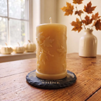 A yellow wax pillar candle with embossed leaf details sits on a dark slate coaster on a wooden table, with a window, pumpkins, and a vase of autumn leaves in the background.