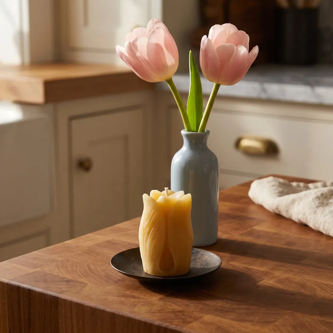 Honey-gold beeswax tulip candle on a black iron dish beside pink tulips on a sunlit butcher block kitchen island.